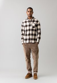 Camicia flanella a quadri beige e nera con bottoni, tasca frontale e maniche lunghe, abbinata a pantaloni di velluto a coste marrone chiaro e scarpe marroni.