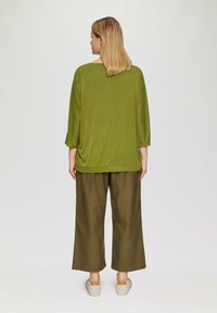 Vrouw met schouderlang blond haar staat met de rug naar de camera, gekleed in een losse groene top, brede olijfgroene broek en witte sneakers.