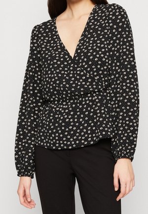 Zwarte wikkelblouse met lange mouwen en klein wit bloemmotief, gedragen met zwarte broek, toont bovenlichaam en handen.