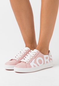 Rosa lädersneakers med vita accenter, med stor "KORS"-logotyp, vita snören och en texturerad gummisula. Lågprofil design.