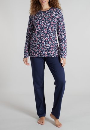 Frau trägt ein marineblaues, langärmliges Oberteil mit Blumenmuster und einfarbige marineblaue Hose, steht barfuß vor einem schlichten grauen Hintergrund.