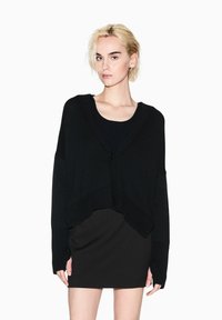 Cardigan nero oversize con scollo a V profondo, texture a costine e chiusura frontale con bottoni, abbinato a un vestito nero aderente.