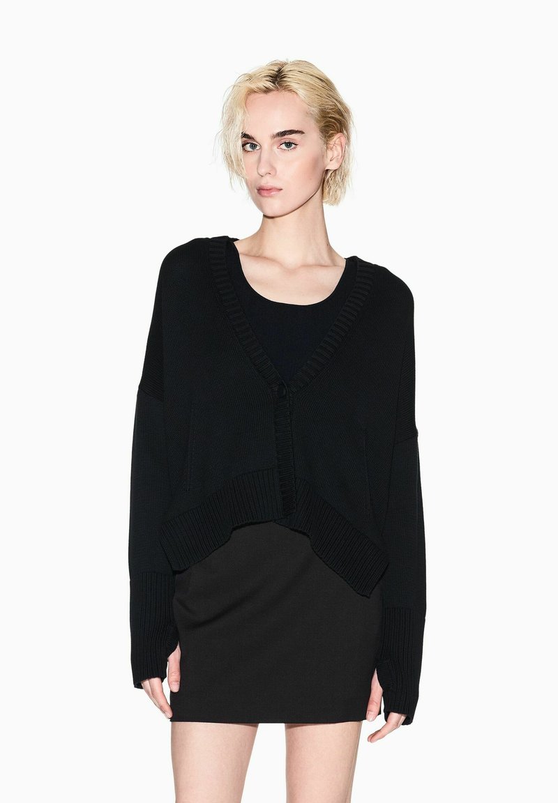 Cardigan nero oversize con scollo a V profondo, texture a costine e chiusura frontale con bottoni, abbinato a un vestito nero aderente.