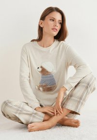 Top de manga larga beige con un oso bordado que lleva un suéter azul y gris, combinado con pantalones de cuadros en colores suaves, sobre una superficie texturizada.