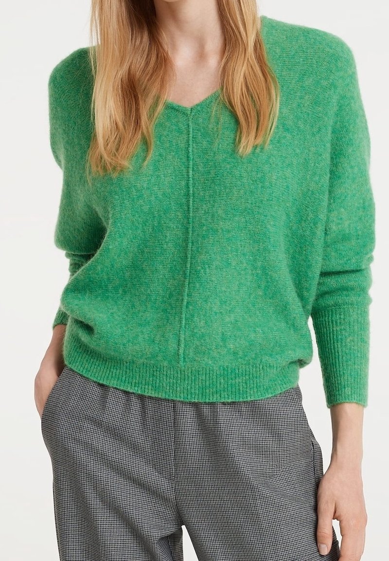 Femme portant un pull vert à col en V rentré dans un pantalon gris à carreaux taille haute, avec une main dans la poche, sur fond blanc.