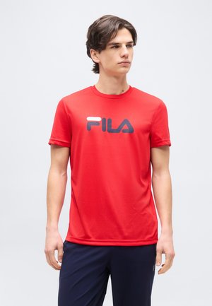 Fiatal férfi piros FILA pólóban és tengerészkék nadrágban, egyszínű világosszürke háttér előtt áll, oldalra néz.