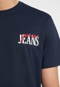 Navyblauwe katoenen T-shirt met witte "JEANS" tekst en een rode cursieve "guess" accent. Korte mouwen en ronde halslijn, gladde textuur.