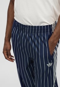 Άνδρας που φοράει σκούρο μπλε φόρμα Adidas με λευκές κάθετες ρίγες και λευκό μπλουζάκι, με το ένα χέρι στην τσέπη, μπροστά σε απλό φόντο.
