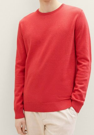 Mężczyzna ubrany w prosty czerwony dziany sweter z prążkowanymi mankietami i dołem, w połączeniu z jasnymi spodniami, stojący na tle jasnej ściany.