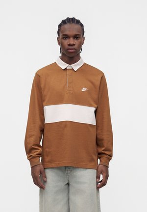 Ung mand med flettet hår iført brun og hvid Nike langærmet polo og lysegrå bukser, stående foran en ensfarvet hvid baggrund.
