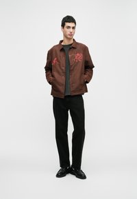 Jovem vestido com um casaco castanho abotoado com bordados vermelhos, calças pretas e sapatos pretos, de pé com as mãos nos bolsos contra um fundo branco.