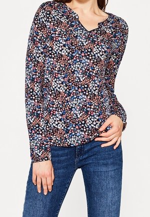 Kobieta w długim rękawie i bluzce z dekoltem w serek, z nadrukiem wielokolorowych kwiatów, w połączeniu z niebieskimi dżinsami, z ręką opartą na biodrze.