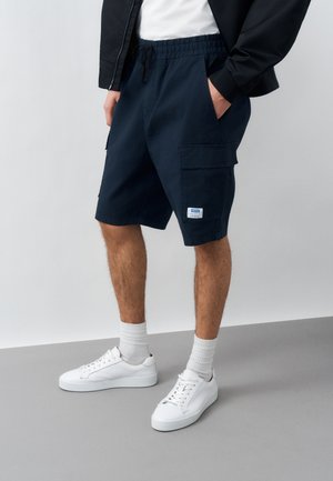 Άνδρας που φοράει ναυτικό μπλε cargo σορτς, λευκές κάλτσες και λευκά αθλητικά παπούτσια, στέκεται με τα χέρια στις τσέπες μπροστά σε γκρι φόντο.