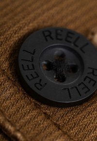 Black plastic button featuring engraved "REELL," set against a textured brown fabric background with visible stitching.