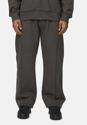 Persona che indossa pantaloni grigi scuri a vestibilità larga con cuciture visibili, felpa con zip abbinata e scarpe da ginnastica nere, in piedi su uno sfondo neutro.