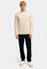 Beiger gestrickter Pullover mit einem strukturierten Rautenmuster, rundem Ausschnitt und gerippten Bündchen, kombiniert mit dunkler Hose und weißen Turnschuhen.