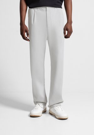 Homme portant un pantalon droit gris clair et des baskets blanches, debout devant un fond blanc uni.