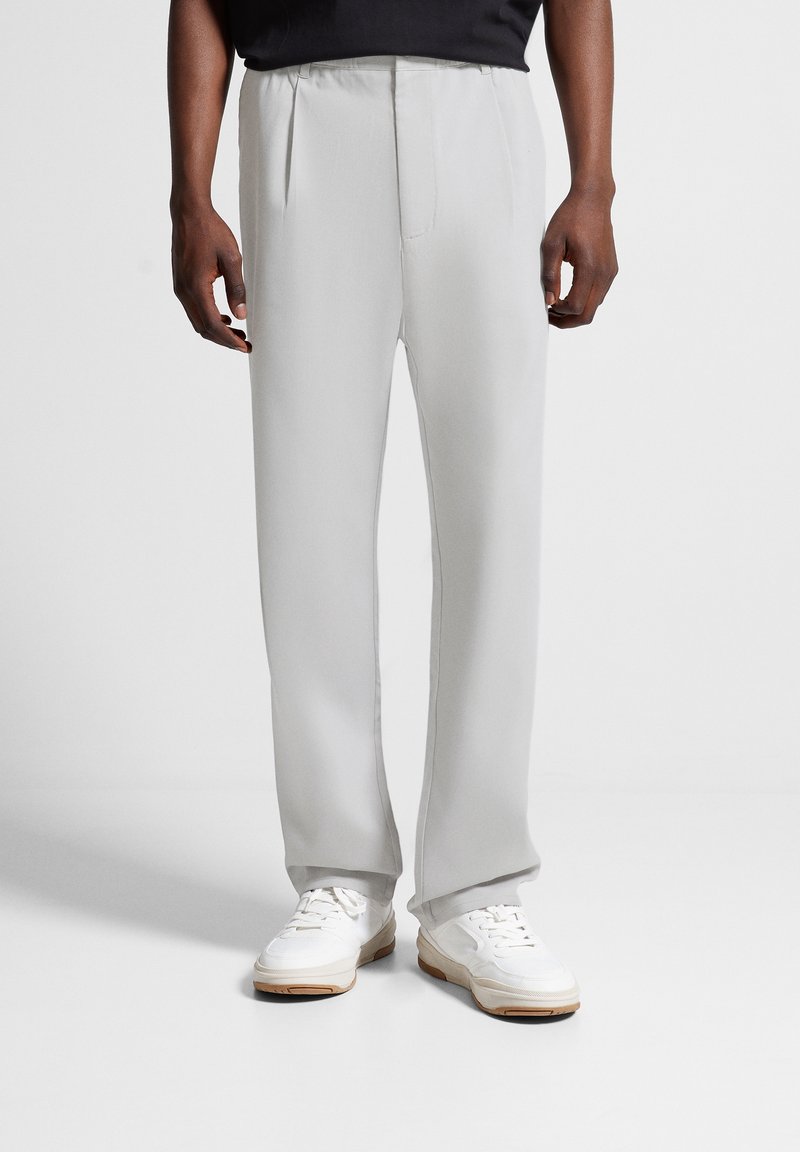 Homme portant un pantalon droit gris clair et des baskets blanches, debout devant un fond blanc uni.