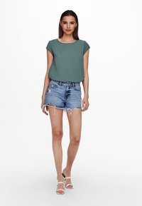 Grünes Kurzarmoberteil kombiniert mit blauen Denim-Shorts. Das Outfit hat einen lässigen Schnitt und fransige Kanten an den Shorts.