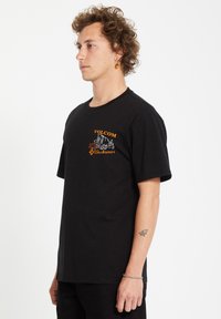 T-shirt en coton noir, à manches courtes, col rond, avec un logo brodé orange sur la poitrine, coupe décontractée, texture lisse.