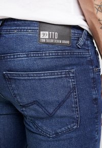 Jeans de mezclilla azul oscuro con un bolsillo trasero que cuenta con un patrón de costura en forma de chevron. La etiqueta trasera dice "TTD Tom Tailor Denim Brand."