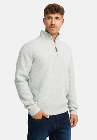 Mand med kort, krøllet hår iført en lysegrå ribbet kvart-zip sweater og mørkeblå jeans, stående med den ene hånd i lommen.