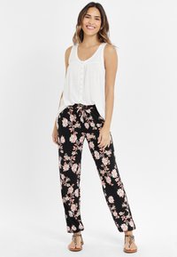 Top bianco senza maniche con bottoni frontali, abbinato a pantaloni neri con motivo floreale caratterizzati da fiori rosa e bianchi. Sandali con dettagli decorativi.