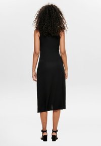 Femme aux cheveux bouclés portant une robe midi noire sans manches et des sandales à talons noires, debout de dos sur un fond clair uni.