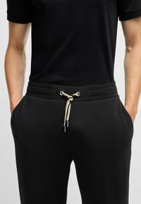 Pantalons de jogging noirs avec une taille côtelée, dotés d'un cordon de serrage avec une rayure beige et blanche, de poches latérales et d'une texture lisse.