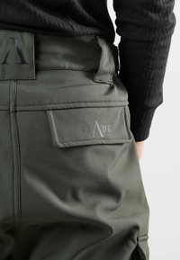 Olijfgroene cargo-broeken hebben een gladde textuur, dubbele zijzakken en een opvallende achterpatch met het merkembleem "SLADE" in grijs.