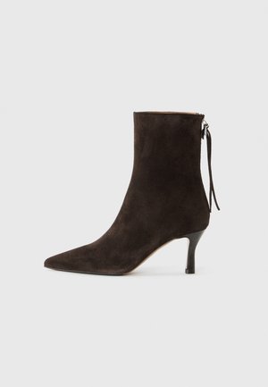 Bottine en daim marron avec un bout pointu et un talon stiletto de hauteur intermédiaire. Présente une fermeture éclair et un détail décoratif de frange.