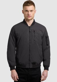 Homme portant une veste bomber noire avec des poches zippées sur la poitrine et les manches, debout devant un fond clair uni.