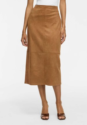 Femme portant une jupe midi taille haute couleur beige associée à des talons ouverts marron sur fond blanc uni.