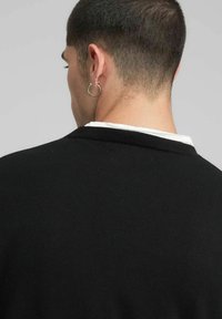 Pull noir avec texture côtelée, col rond, et une couche blanche visible au niveau du col. Le modèle porte une boucle d'oreille argentée en forme d'anneau.
