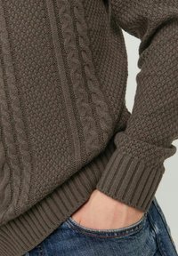 Ciemnobrązowy sweter z dzianiny, z teksturowanymi wzorami, splotem warkoczowym, ściągaczami na mankietach i w pasie. Zestawiony z dżinsami noszonymi w talii.