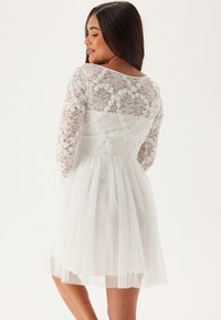 Robe en dentelle blanche avec des manches longues, présentant un corsage ajusté et une jupe évasée et transparente. Détails en dentelle florale sur les manches et le dos.