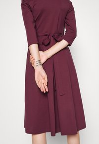 Robe mauve mi-longue à manches trois quarts, ceinturée à la taille. Tissu doux avec une texture lisse et un design simple.