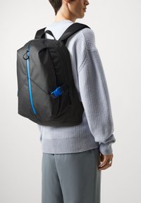 Svart ryggsäck med en glänsande yta, med blå accenter och en geometrisk design, bärs på en persons rygg mot en enfärgad bakgrund.