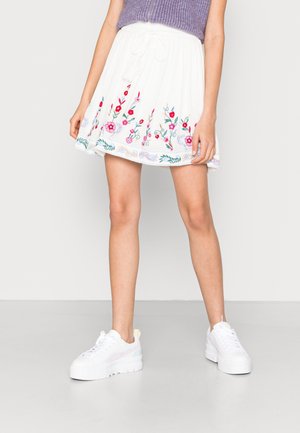 Person iført en hvid mininederdel med farverig blomsterbroderi, en lilla ærmeløs top og hvide platform-sneakers, stående på en grå gulv.