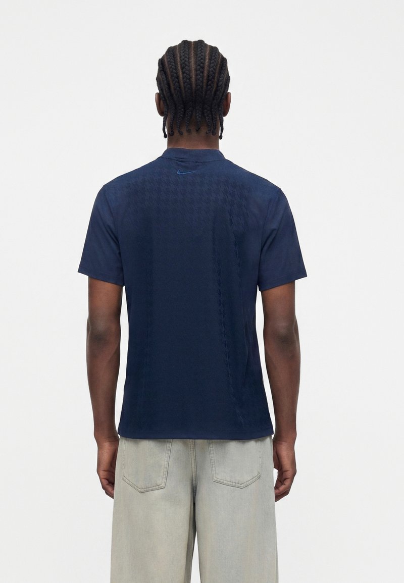 Man with braided hair wearing a navy patterned Nike polo shirt and light gray pants, shown from the back on a white background.