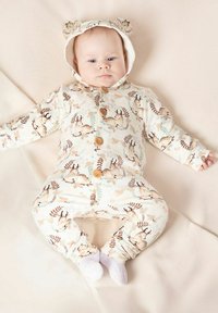 Kremowy kombinezon z kapturem z uszkami niedźwiedzia, drewnianymi guzikami oraz nadrukami nawiązującymi do przyrody, w tym sarną i liśćmi. Miękki, elastyczny materiał.