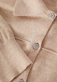 Cardigan beige realizzato in morbido materiale a maglia, con colletto classico, chiusura a bottoni e tessuto testurizzato con una leggera lucentezza.