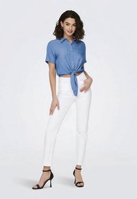 Γυναίκα με σγουρά σκούρα μαλλιά, φορεί μπλε κοντομάνικη μπλούζα δεμένη, άσπρο παντελόνι ψηλής μέσης και μαύρες ψηλοτάκουνες σάντες, στέκεται.