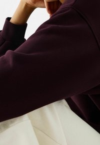 Persona descansando la barbilla en la mano, vistiendo una camiseta de manga larga morada oscura y pantalones de color claro, contra un fondo blanco simple.