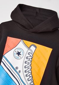 Černá mikina s kapucí, která má grafický potisk tenisky Converse v modré a oranžové barvě, s texturovaným písmem a kruhovým logem.