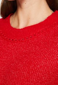 Pull en tricot rouge avec un tissu texturé, un col rond et un motif de point décoratif le long des bords du col. Apparence douce et chaleureuse.