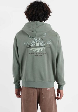 Sweat à capuche vert avec un dessin graphique représentant un soleil et des vagues dans le dos. Le tissu semble doux avec une poche kangourou. Pantalon marron.