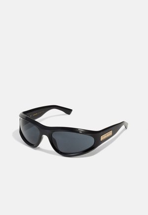 Lunettes de soleil noires avec une monture en plastique brillant, design enveloppant, verres teintés foncés et détail du logo doré sur la branche.