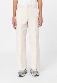 Pantalones de tiro ancho en color beige con una textura suave, que cuentan con paneles cosidos y una etiqueta de marca visible en el lateral, combinados con zapatillas gris.