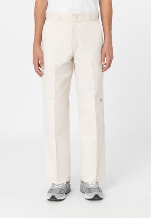 Pantaloni beige a gamba larga con una texture liscia, dotati di pannelli cuciti e un'etichetta del marchio visibile di lato, abbinati a sneakers grigie.